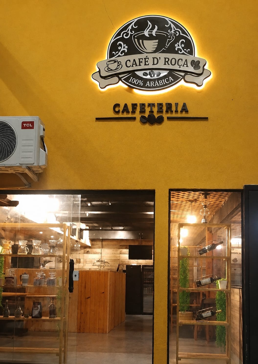Fachada completa da Café D' Roça: placa iluminada na parede amarela e portas de entrada